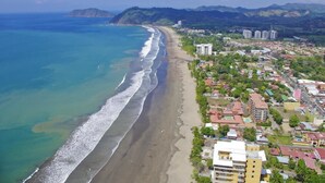 An aerial view of a coastal town with a beach, buildings, and a mountainous backdrop. - Club del cielo Unit 1 (Jacó)