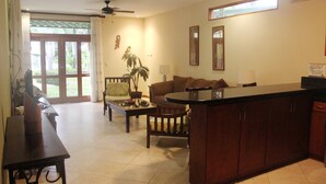 A spacious living area with a sofa, coffee table, and a dining table. There is a ceiling fan, a large window with curtains, and a kitchen area with cabinets. - Club del cielo Unit 1 (Jacó)