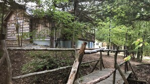 Un puente de madera rústico en un área boscosa, con una cabaña rústica al fondo.