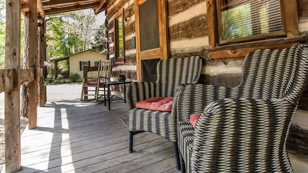 Una cabaña de madera con porche, mobiliario de mimbre y vistas del bosque circundante.