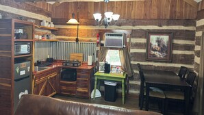 Interior rústico de una cabaña con una mesa de comedor de madera, una mesa verde, una kitchenette con microondas, estufa y pileta. Hay un cuadro en la pared y un ventilador de techo.