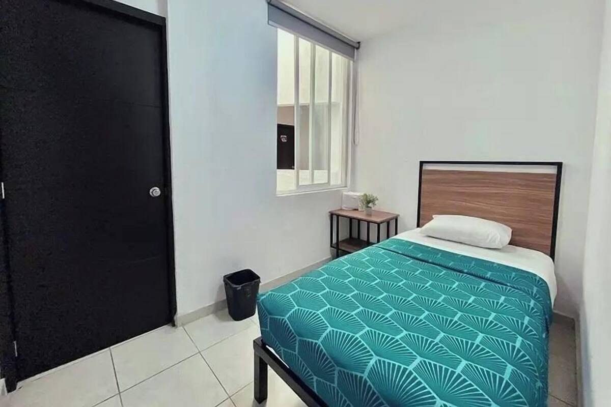 Un dormitorio con cama, ventana con persianas, mesita de noche pequeña y un cubo de basura.