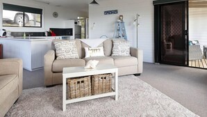A cozy living room with a sofa, a coffee table, and a kitchen area in the background. - KINGFISHER COTTAGE Beachfront at Bay of Fires (Bay of Fires)