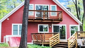Uma cabana vermelha de dois andares, com varanda, deck de madeira e chão de terra.