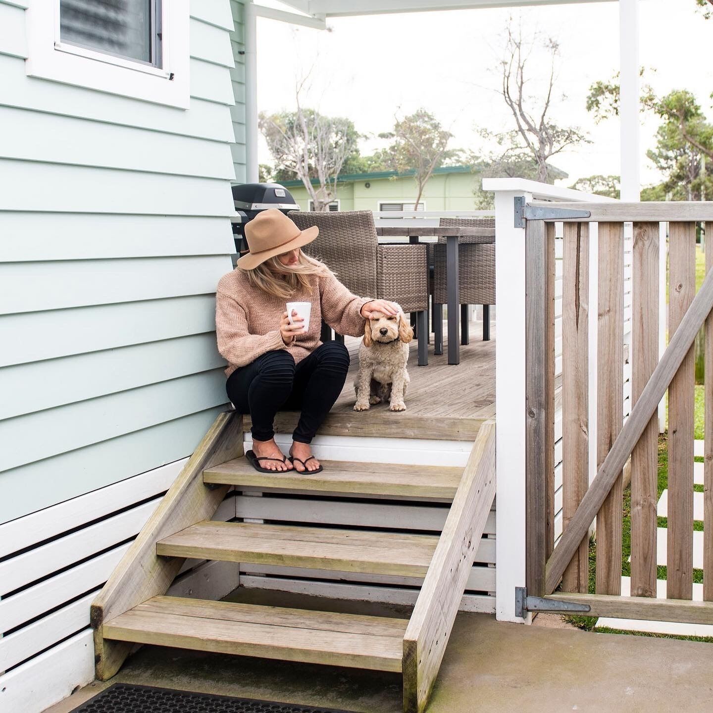Une personne assise sur des marches extérieures, accompagnée d’un chien, une terrasse avec des meubles en osier et une clôture en bois.