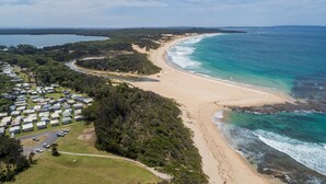 An aerial view of a coastal area with a beach, a campground with RVs, and a dense forest. - Surfside Cudmirrah Beach (Cudmirrah)