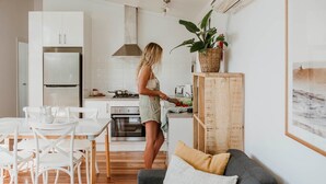 A modern kitchen with white cabinetry, a dining area with white chairs, and a grey sofa with cushions. - Surfside Cudmirrah Beach (Cudmirrah)
