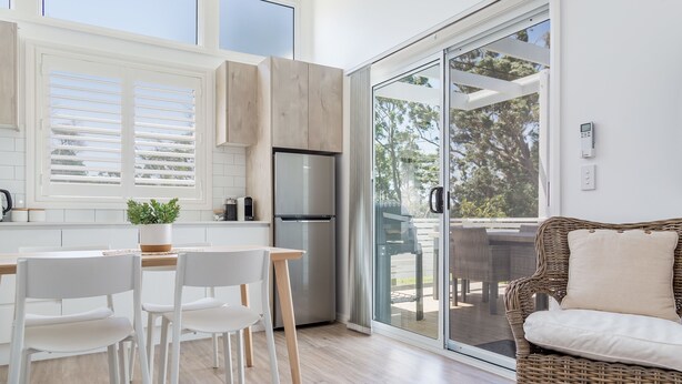A modern kitchen with white cabinetry, a built-in refrigerator, and a dining area with white chairs and a table. - Surfside Cudmirrah Beach (Cudmirrah)