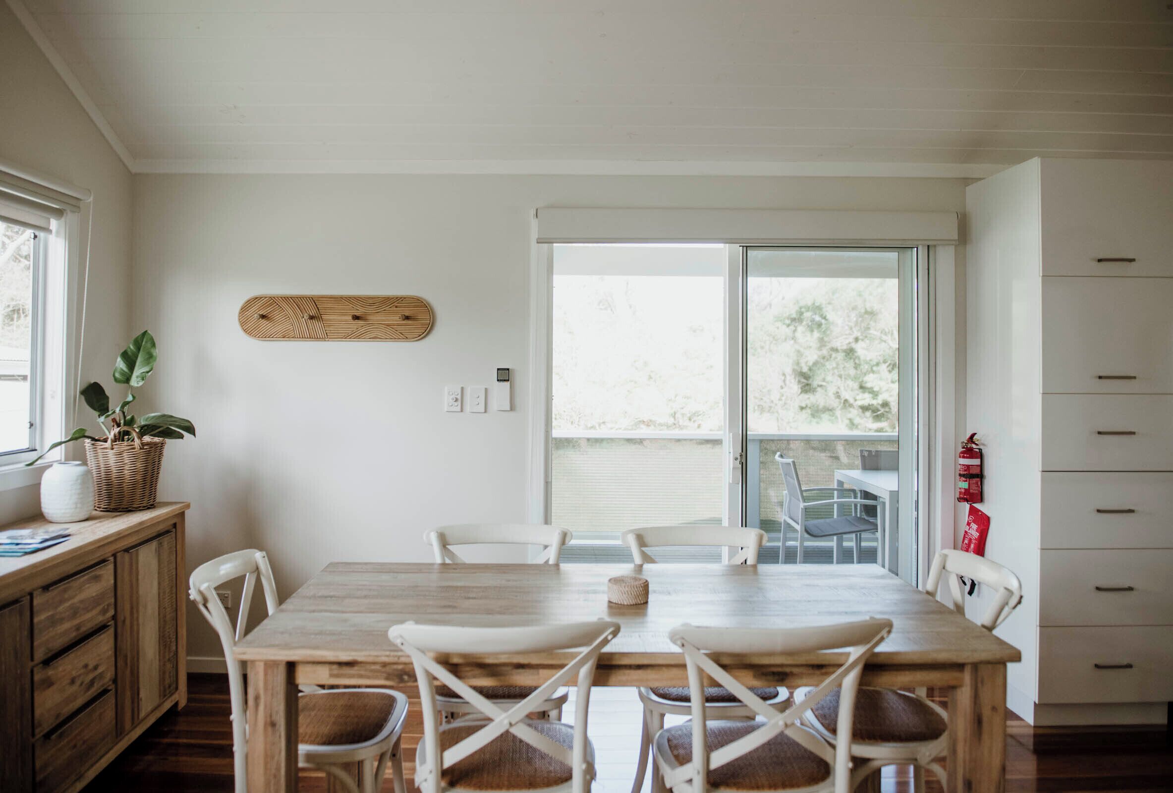 A dining area with a wooden table and white chairs, a basket with a plant, and a window with a view of greenery.