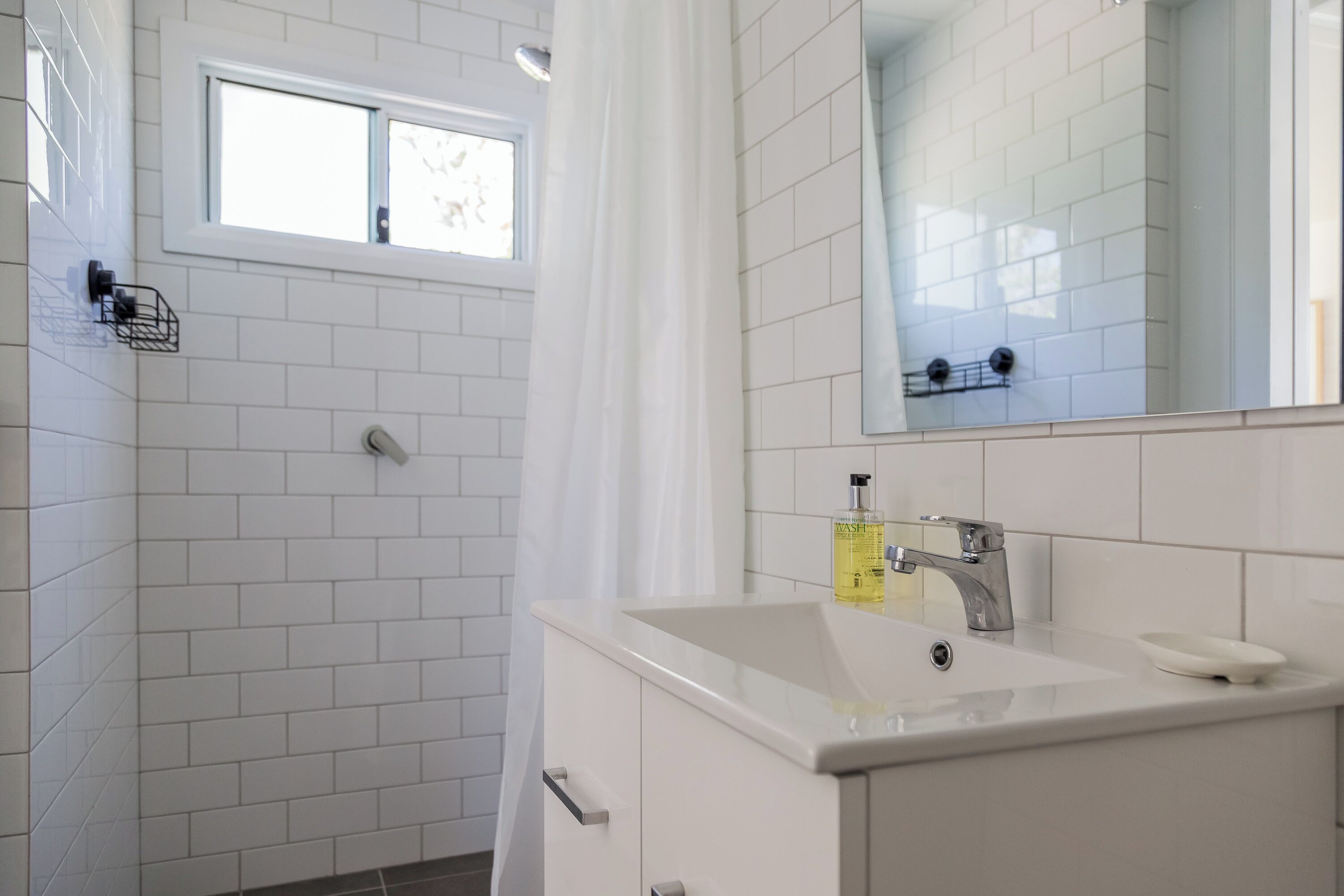 Une salle de bain moderne avec des carreaux blancs, une fenêtre, une douche, un lavabo avec un miroir et un porte-savon.
