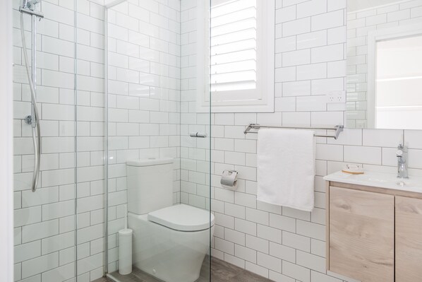 A bathroom with white tiled walls, a glass shower enclosure, a toilet, a sink with a mirror, and a towel rack. - Surfside Cudmirrah Beach (Cudmirrah)