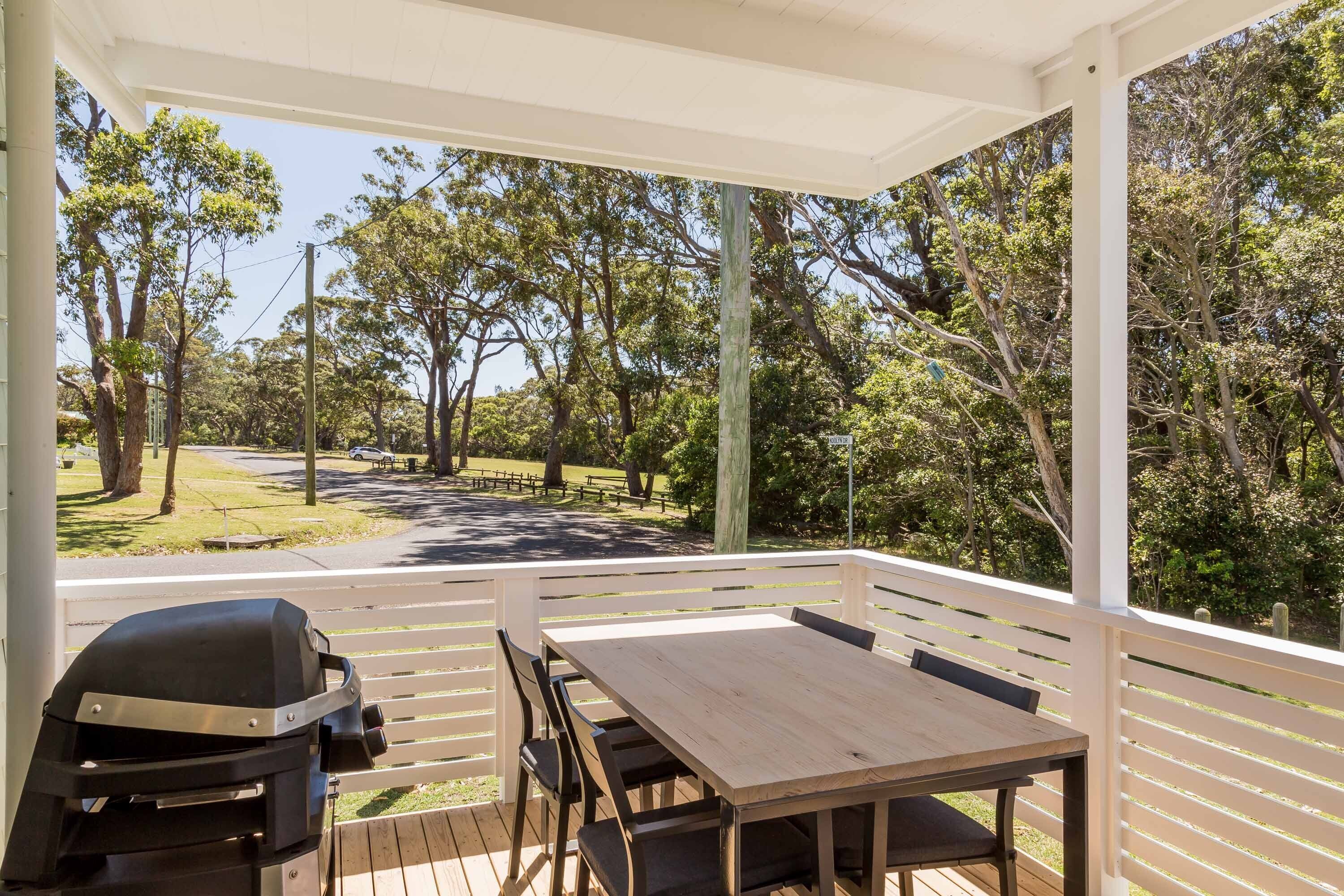 A covered outdoor area with a wooden table and chairs, a barbecue grill, and a view of a park with trees and a pathway.