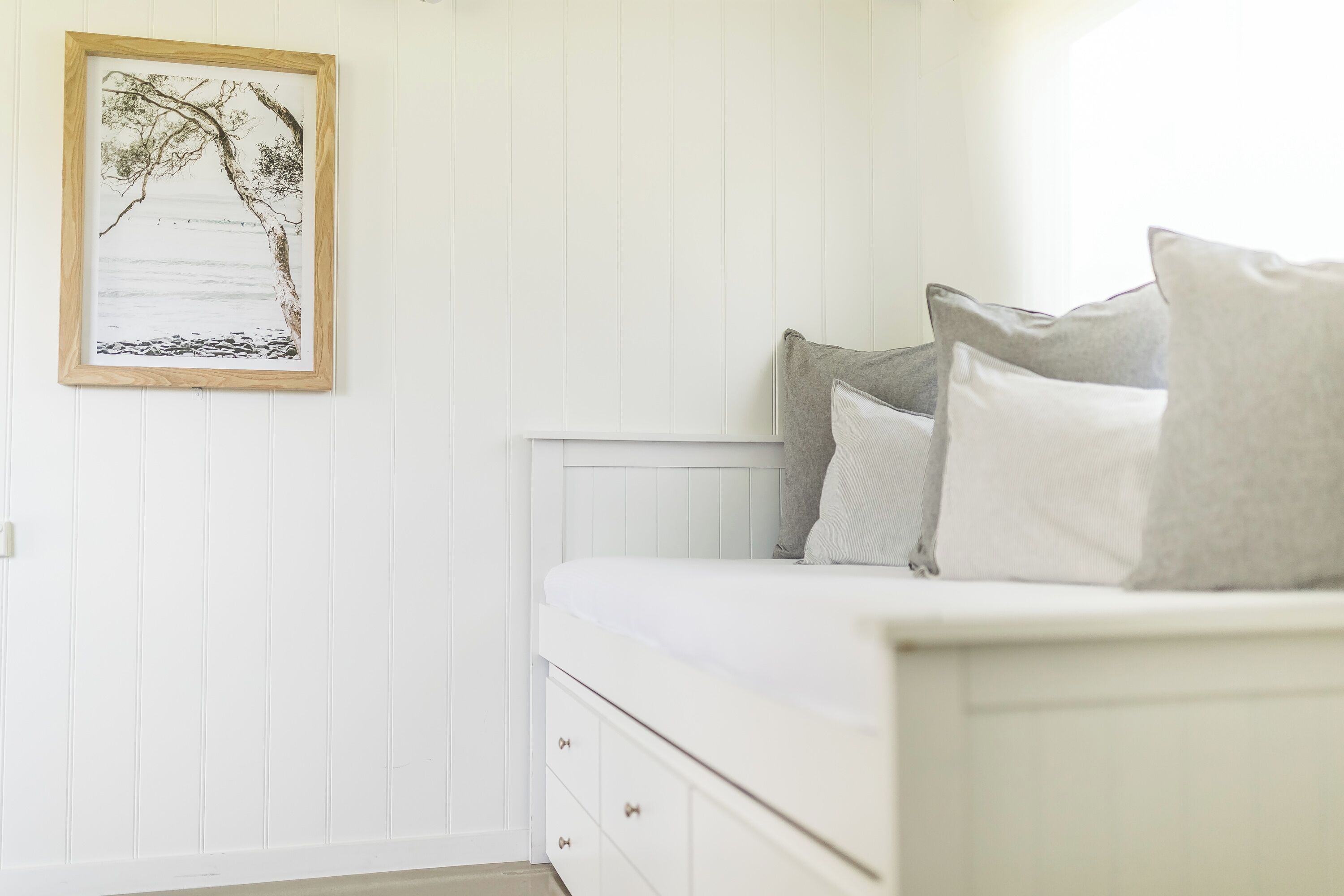 A bedroom with a white bed, grey pillows, and a framed picture on the wall.