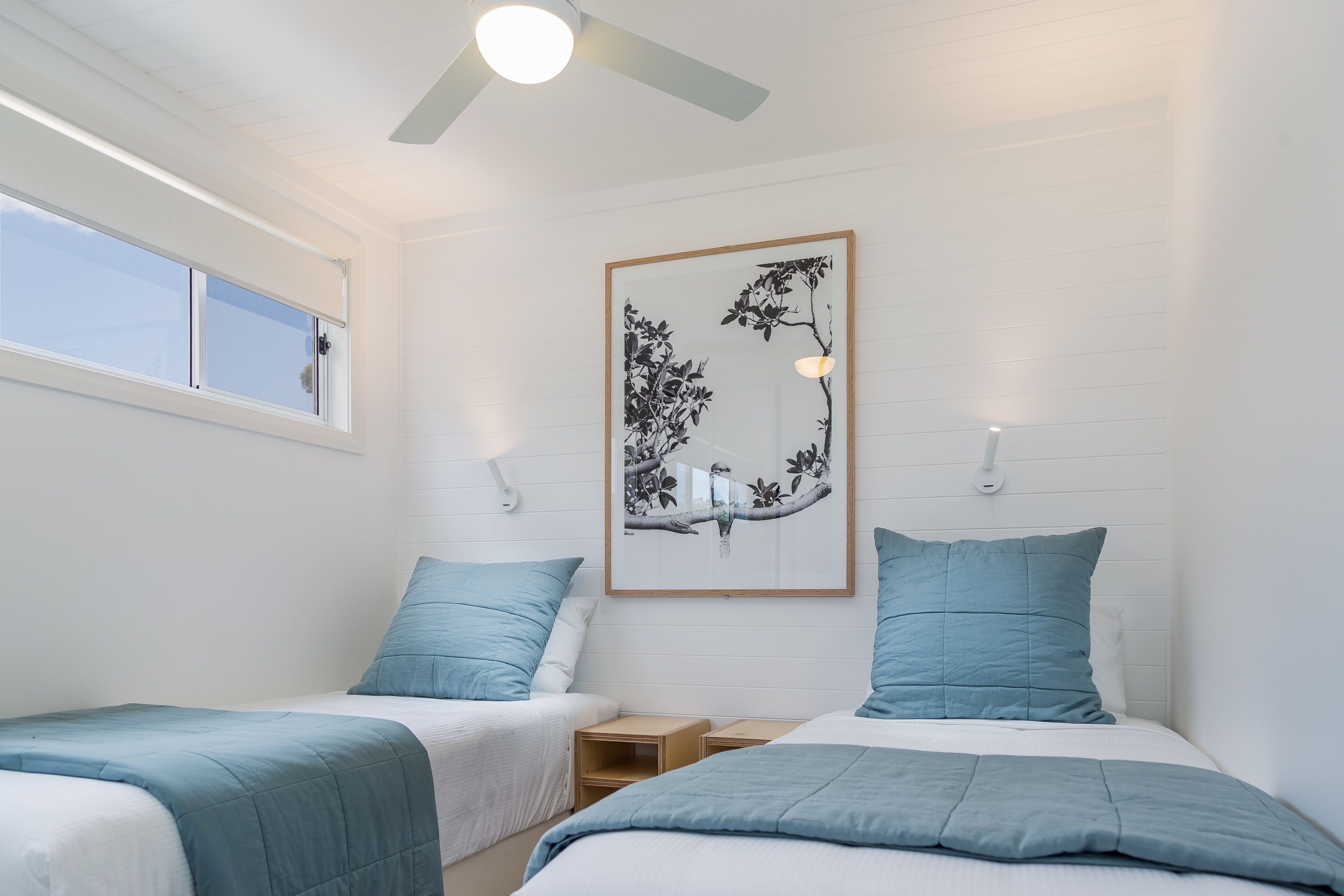 A bedroom with two beds, a ceiling fan, a window, and a wall-mounted artwork.