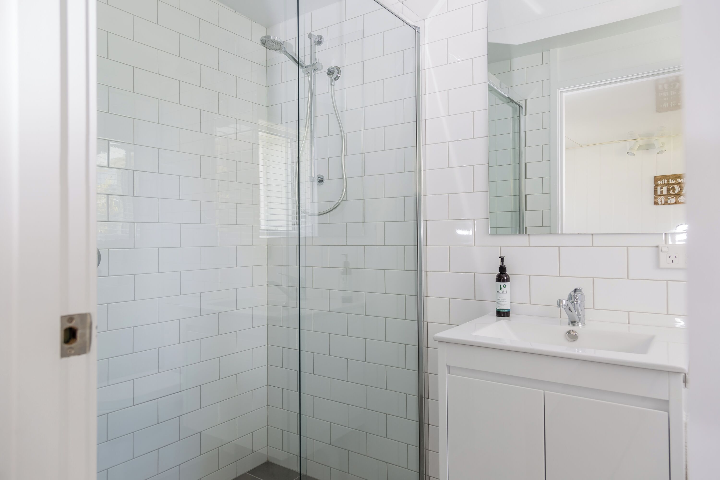 A modern bathroom with white tiled walls, a glass-enclosed shower, and a white sink with a mirror above it.