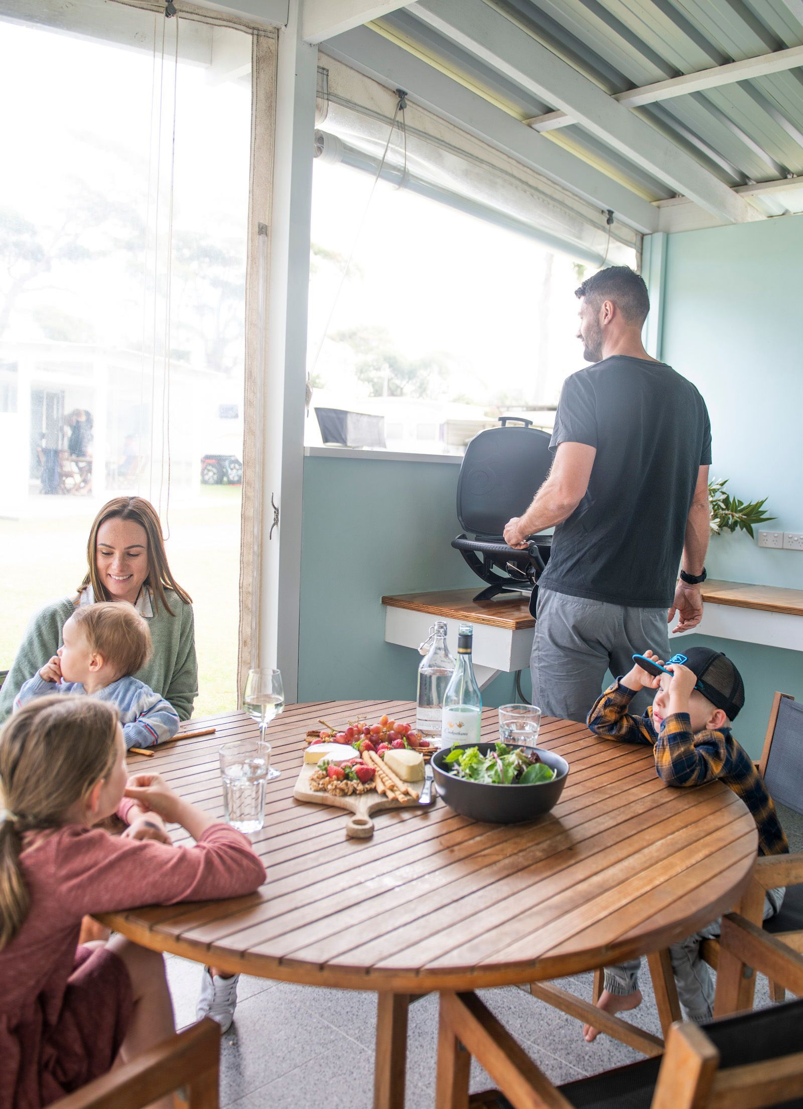 Una familia cenando en una mesa de madera, con comida, bebidas, una parrilla y vistas al exterior.
