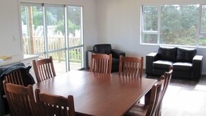 A dining area with a wooden table and chairs, a black sofa, and large windows with a view of greenery.