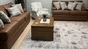 Un salon avec deux canapés marron, une table basse en bois, un tapis à motifs, une horloge murale et une lampe sur une table d’appoint.