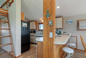A compact kitchen with wooden cabinets, a black refrigerator, a wooden bar counter with stools, and a decorative anchor on the wall.
