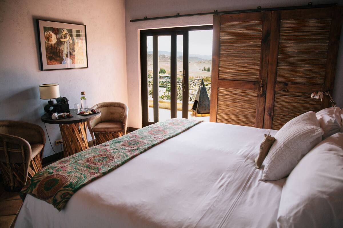 A hotel room with a large bed, a small table with a lamp, a chair, and a view through a sliding door.