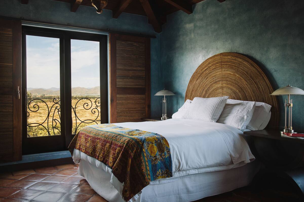 A bedroom with a bed, two bedside lamps, a large window with a view of the countryside, and a wooden ceiling.