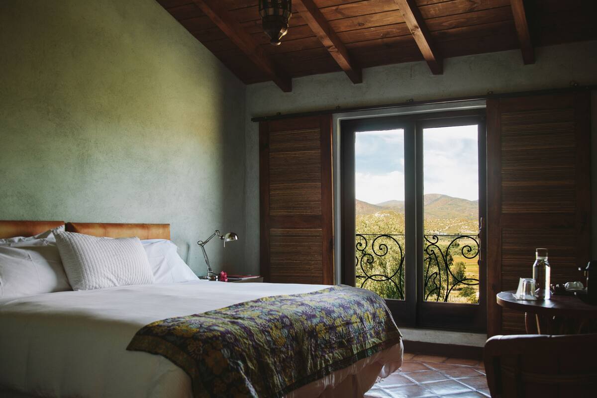 A bedroom with a large bed, a view of the countryside through a window, and a balcony with a wrought iron railing.