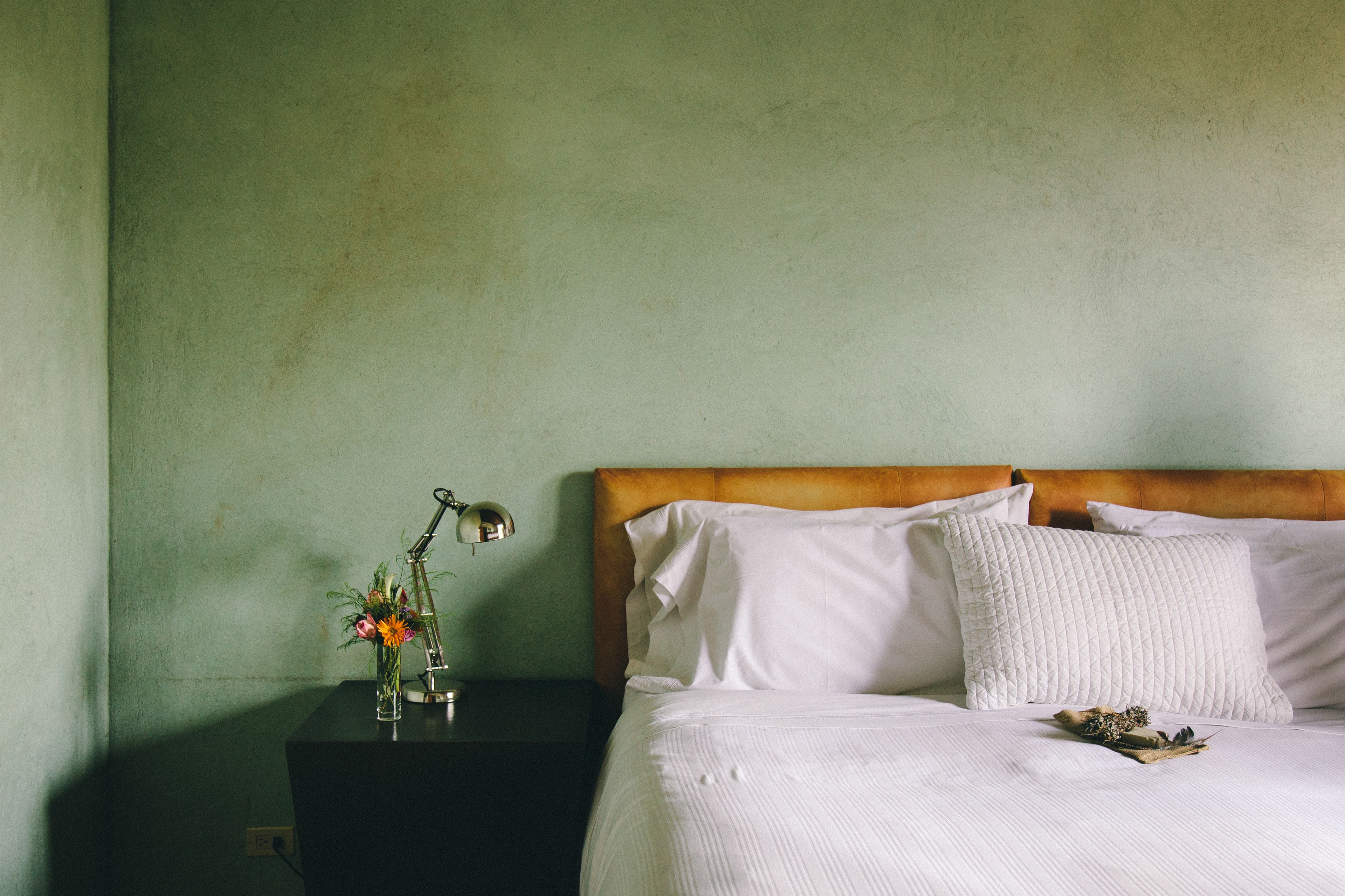 A neatly made bed with white linens and pillows, a bedside table with a lamp and a vase of flowers, and a green wall in the background.
