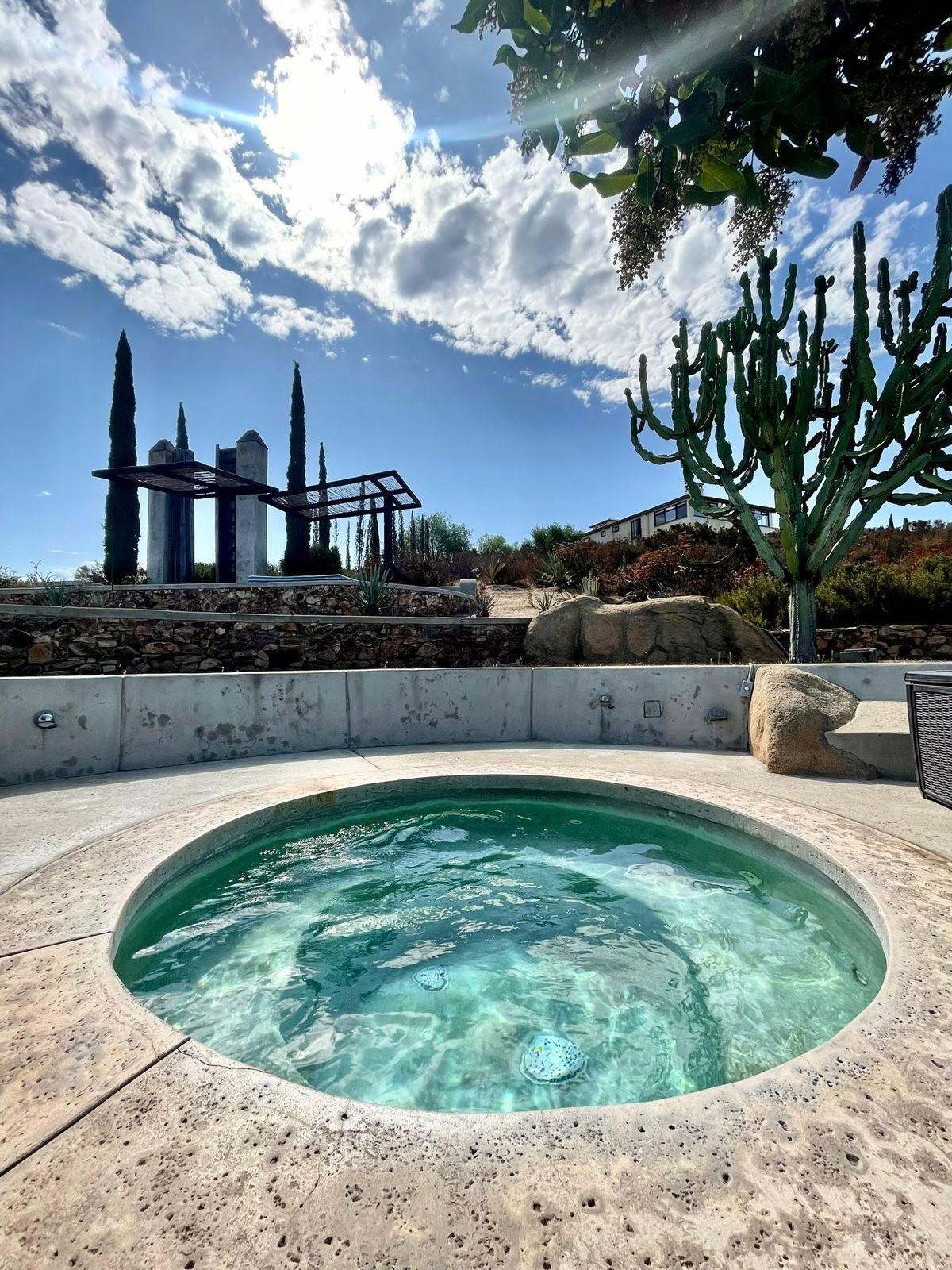 Un jacuzzi rodeado por un muro de piedra, con agua cristalina y vistas a cactus altos y un edificio moderno al fondo.