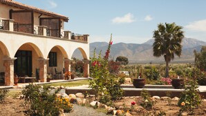 A building with arched windows and a balcony, surrounded by a garden with various plants and flowers, and mountains in the background.