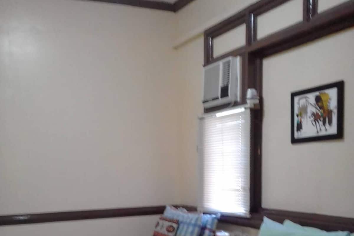 A room with two beds, each with a striped bedspread, a desk with a chair, an air conditioning unit, and a framed picture on the wall.