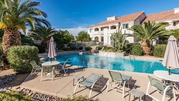 A pool area with lounge chairs and umbrellas, surrounded by palm trees and a building with a tiled roof. - Picture Perfect Paradise Valley Condo (Paradise Valley)