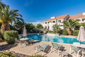 A pool area with lounge chairs and umbrellas, surrounded by palm trees and a building with a tiled roof. - Picture Perfect Paradise Valley Condo (Paradise Valley)