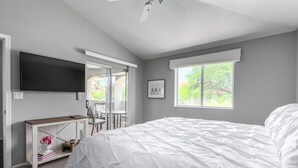 A bedroom with a large bed, a TV mounted on the wall, a desk with a chair, and a window with a view of trees. - Picture Perfect Paradise Valley Condo (Paradise Valley)