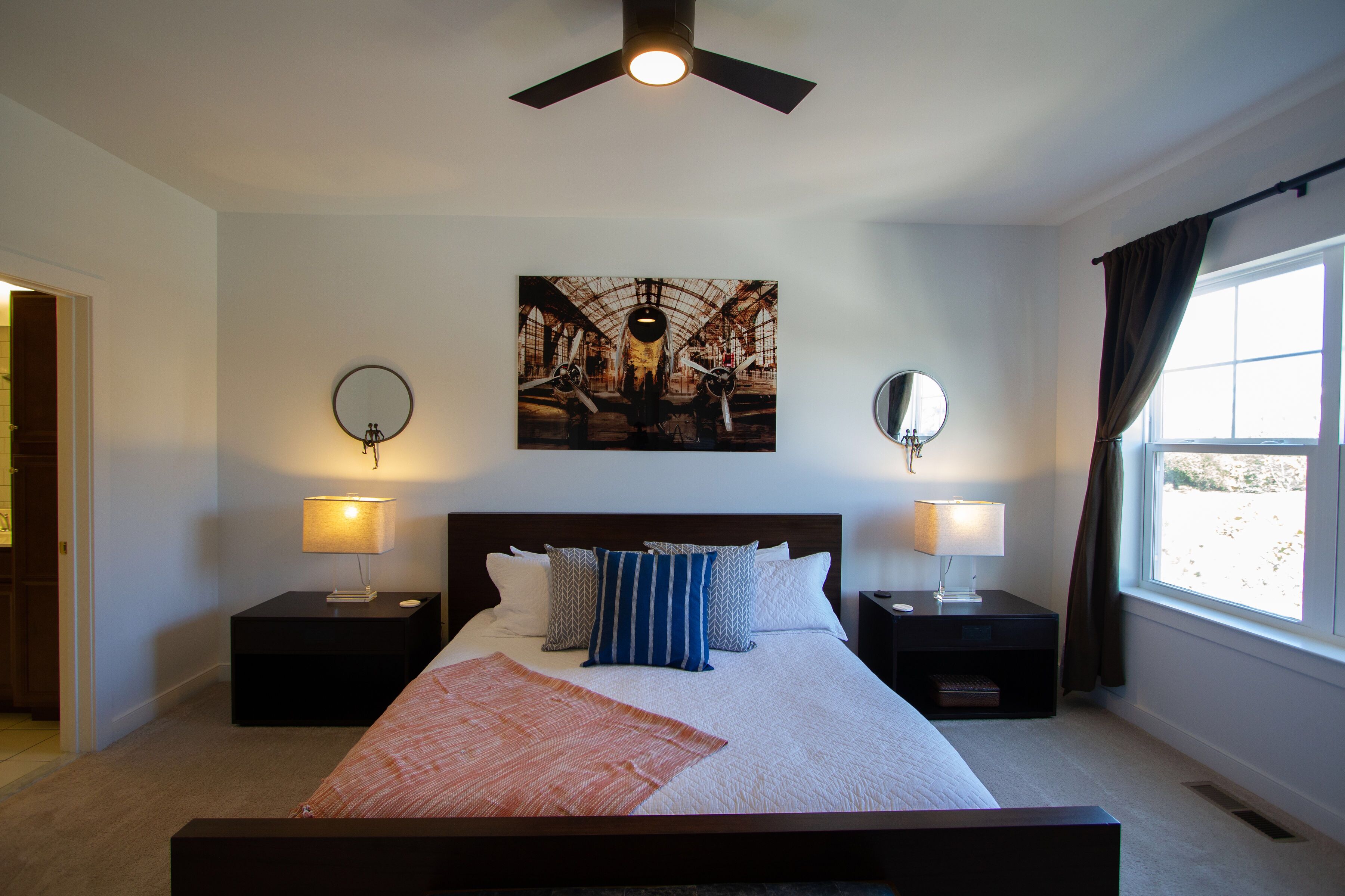 A bedroom with a bed, two bedside tables, two wall-mounted lamps, a ceiling fan, a large window with curtains, and a painting above the bed.