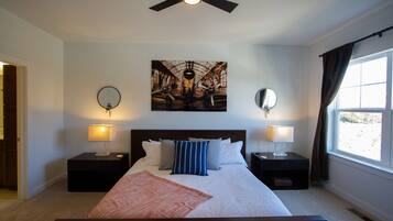 A bedroom with a bed, two bedside tables, two wall-mounted lamps, a ceiling fan, a large window with curtains, and a painting above the bed.