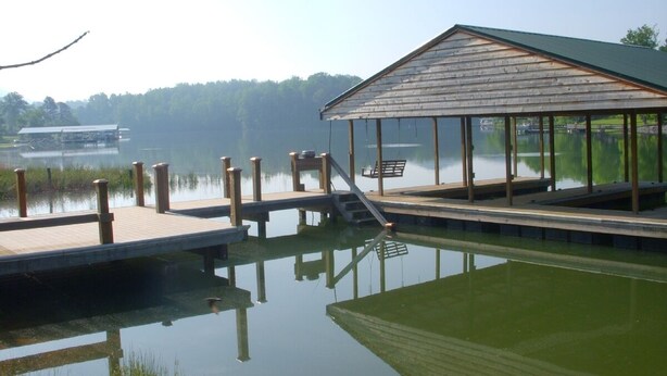 A wooden dock extending into a calm body of water with a covered seating area and a boat docked in the background. - Cozy Waterfront Chalet with Great Dock (HIAWASSEE)