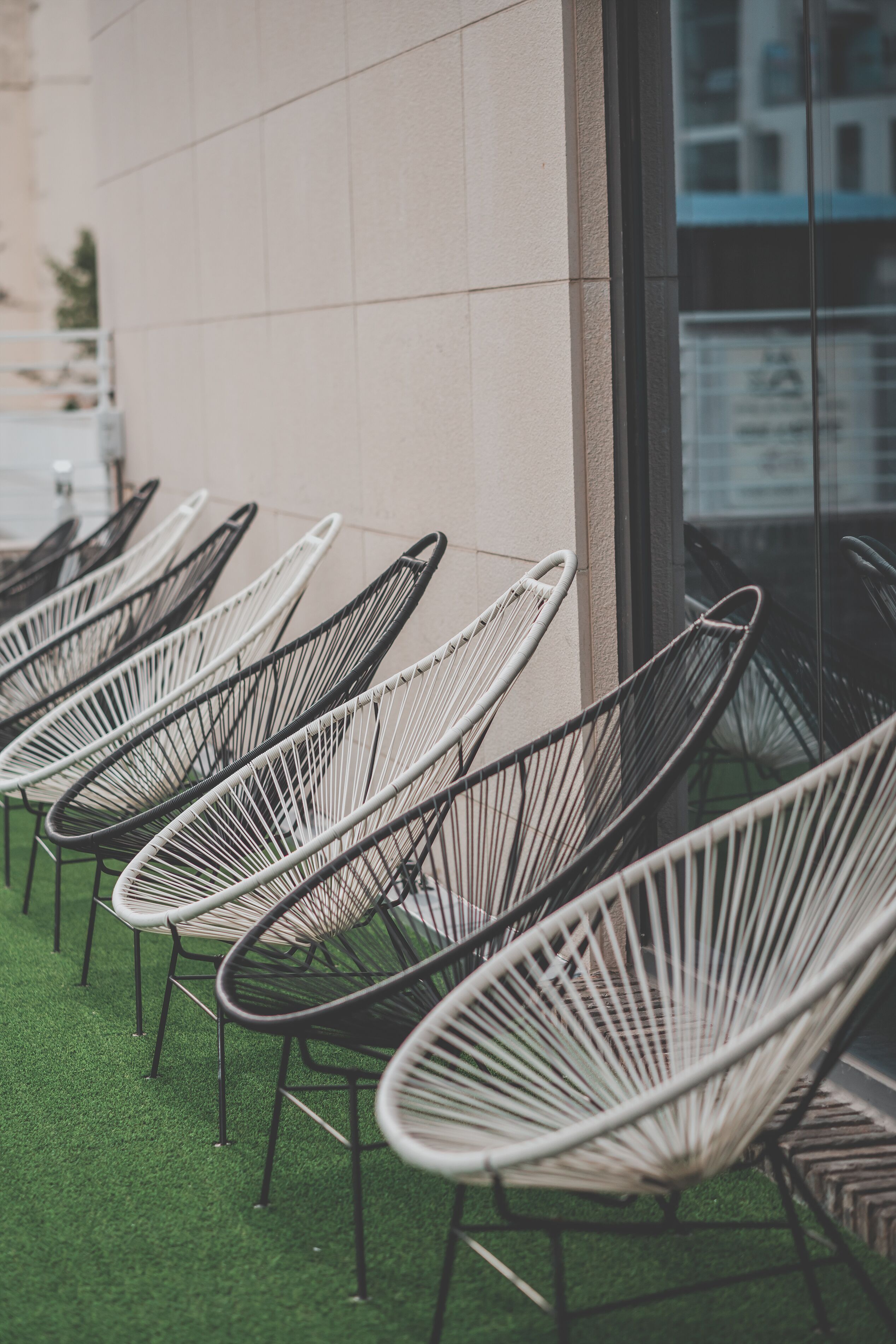 A row of wicker chairs aligned against a building facade with a tiled wall and a glass window.