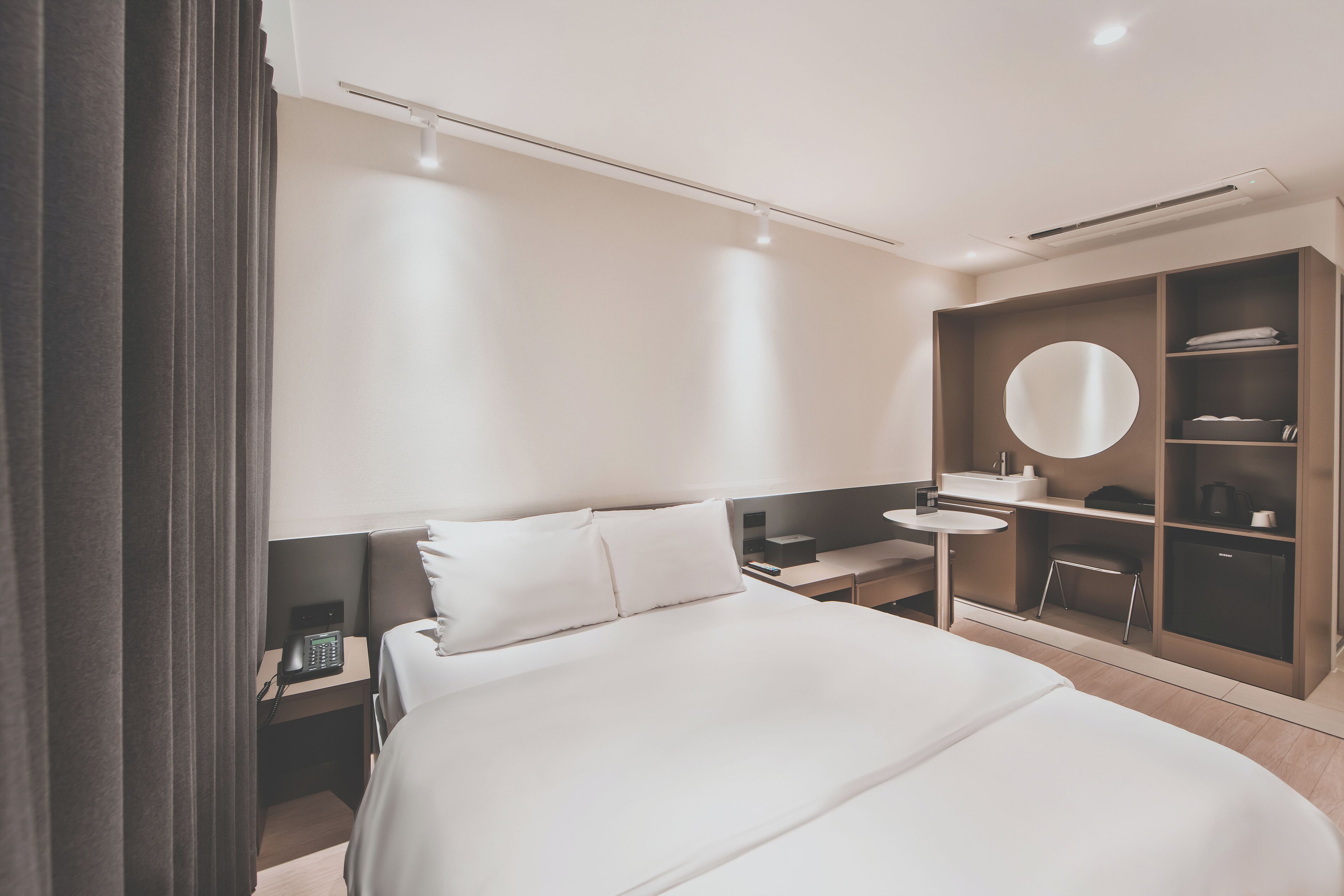 A modern hotel room with a large bed, bedside table, a small desk with a chair, a round mirror, and a wardrobe.