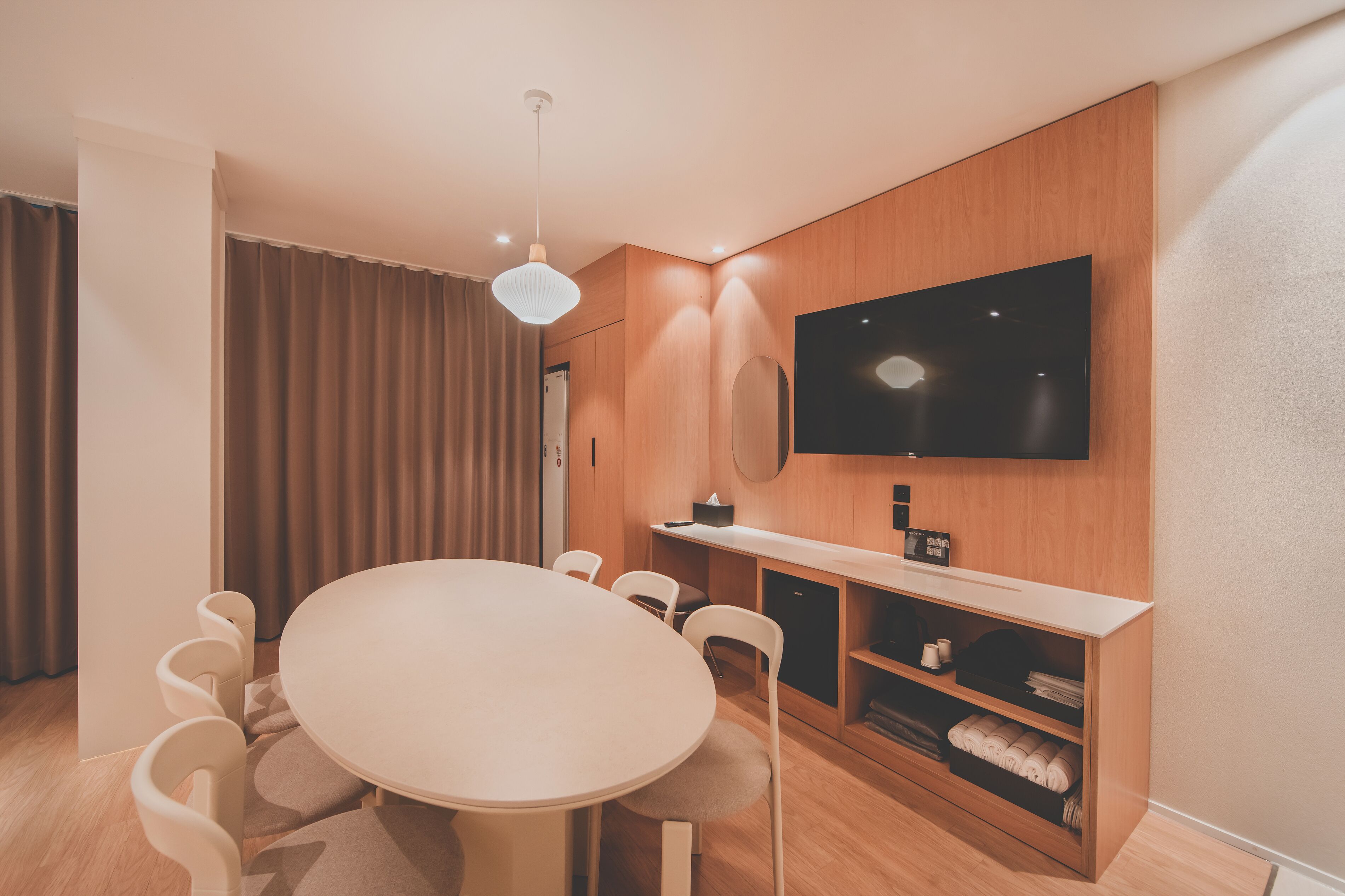 A modern hotel room with a round dining table, white chairs, a flat-screen TV mounted on the wall, and a wooden cabinet with a mirror.