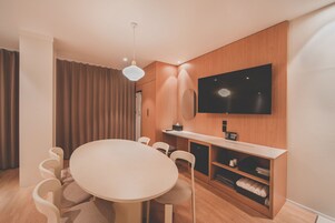 A modern hotel room with a round dining table, white chairs, a flat-screen TV mounted on the wall, and a wooden cabinet with a mirror.