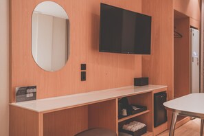Habitación de hotel con televisor de pantalla plana montado en la pared, un espejo redondo, un escritorio blanco y una pequeña zona de estar.