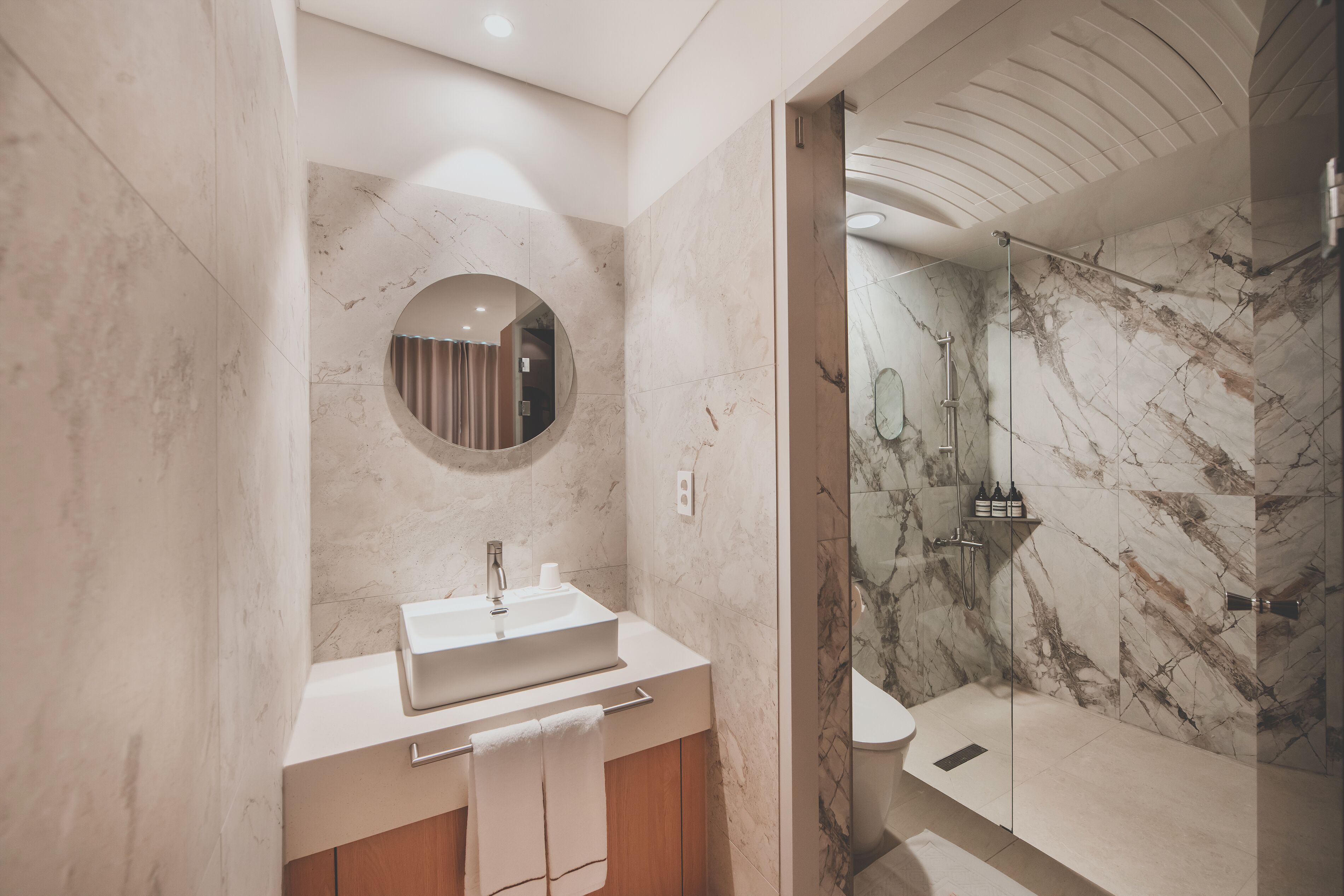 A modern bathroom with a marble sink, a round mirror, and a shower area.