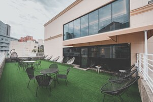 Terraza en la azotea con césped artificial, mobiliario exterior y un edificio con fachada de vidrio.