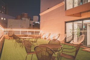 An outdoor seating area with tables and chairs, a glass door leading inside, and a view of the cityscape in the background.