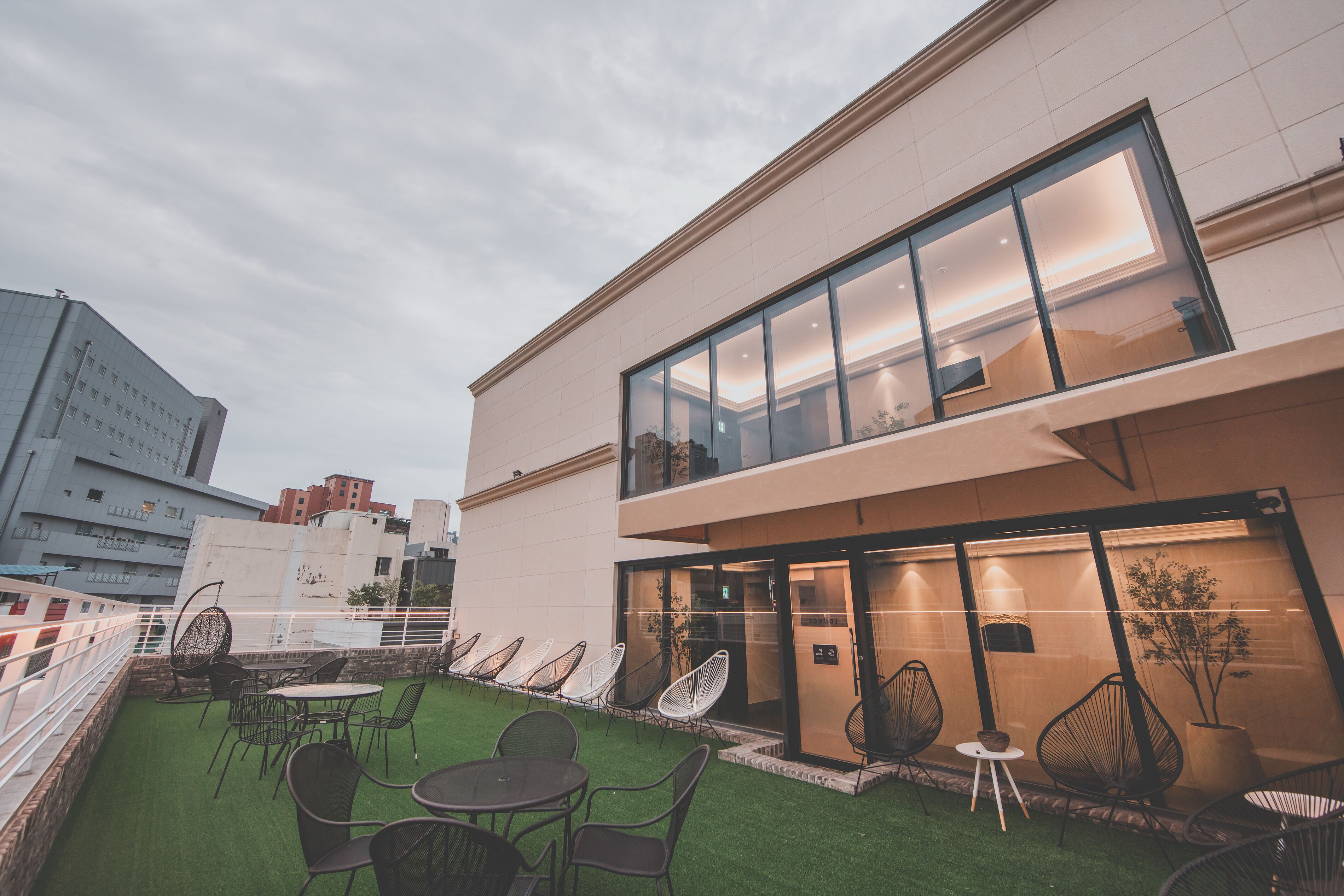 A modern building with a glass facade, outdoor seating area with chairs and tables, and a view of the cityscape in the background.