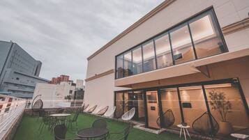 A modern building with a glass facade, outdoor seating area with chairs and tables, and a view of the cityscape in the background.