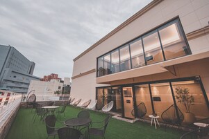 Un edificio moderno con fachada de vidrio, zona de estar al aire libre con sillas y mesas, y vista del paisaje urbano de fondo.