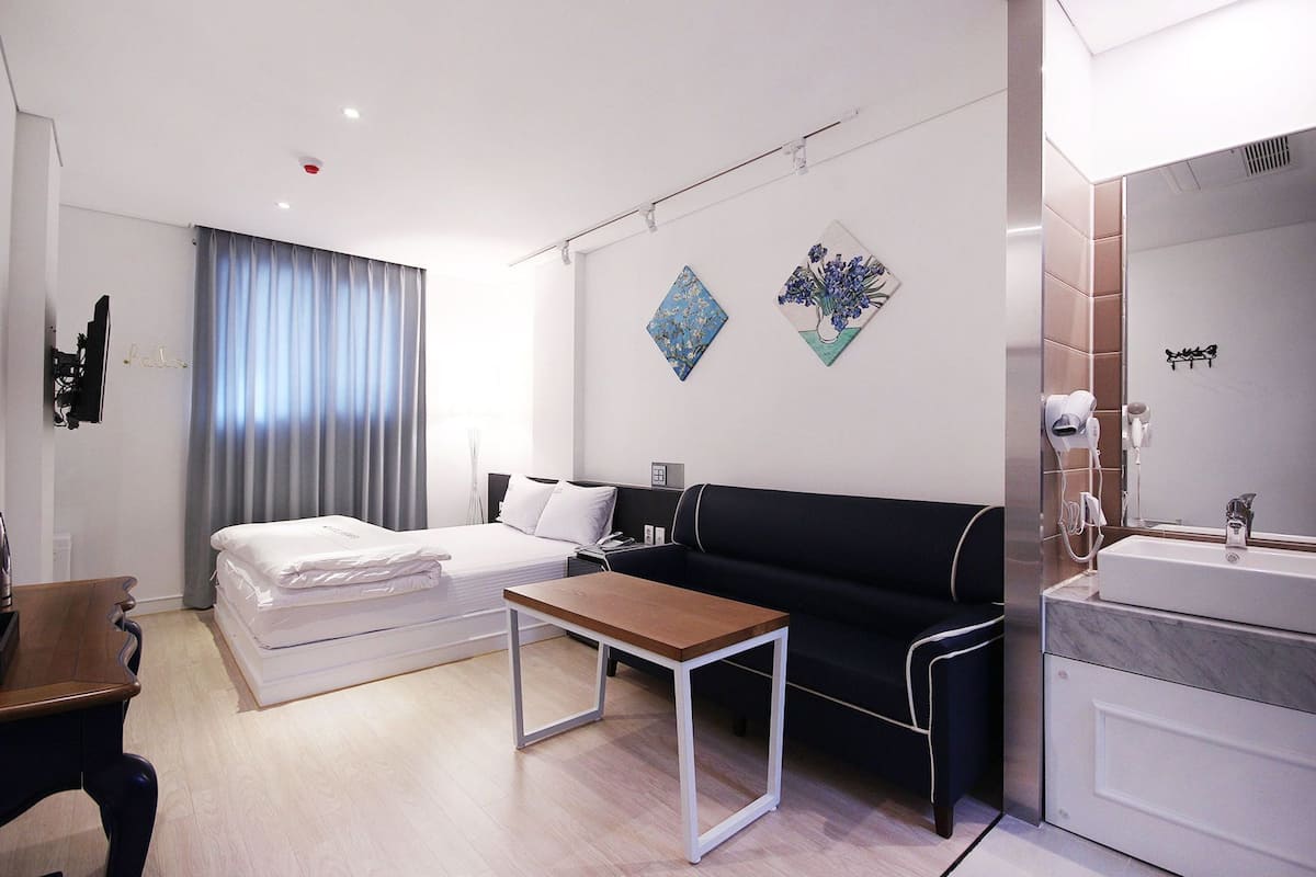 Una habitación de hotel moderna con una cama, un sofá, una mesita, un escritorio, un espejo y un baño con lavamanos y secador de pelo.