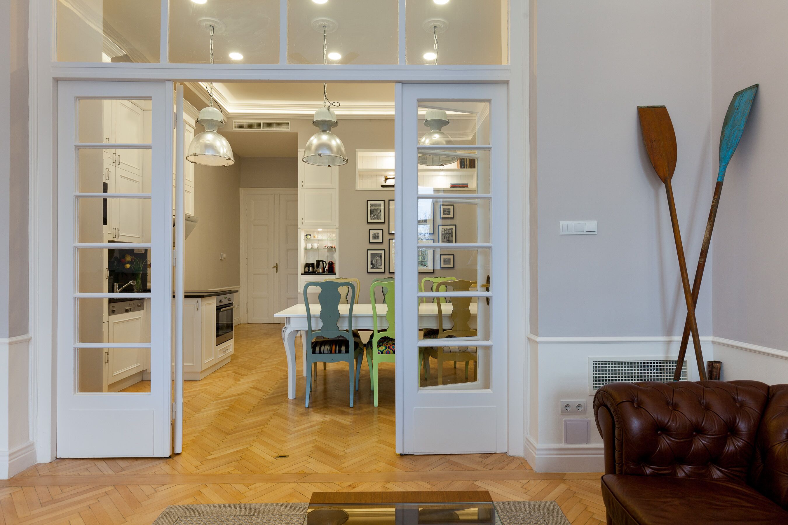 An open door leading to a dining area with a wooden herringbone floor, a dining table with chairs, and a brown leather sofa.