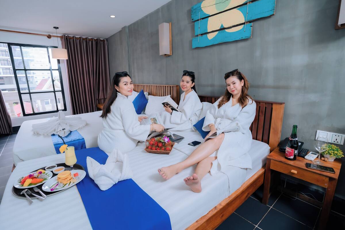 A hotel room with a bed, a bedside table with a bottle, a remote, and a glass of wine. There is a tray with food and drinks on the bed.